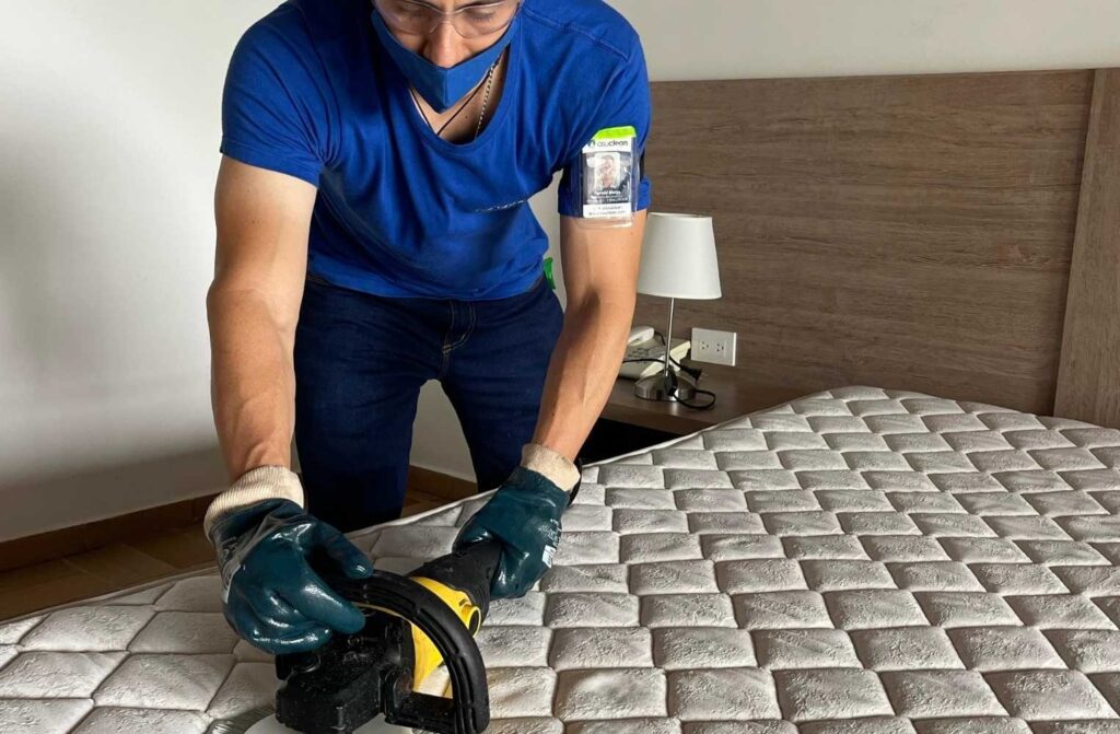 Hombre con camisa azul y guantes usa una aspiradora para limpiar un colchón en Manizales, servicio de limpieza a domicilio.