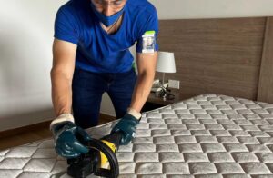 Hombre con camisa azul y guantes usa una aspiradora para limpiar un colchón en Manizales, servicio de limpieza a domicilio.